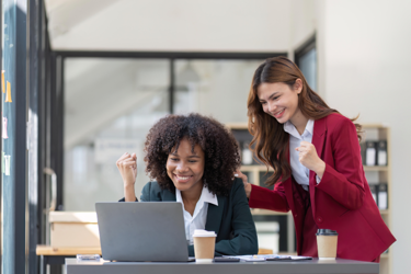 Two people in professional suits at a desk with an open laptop, papers, and two coffee cups