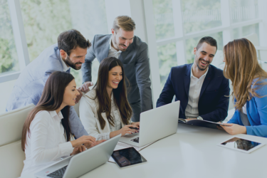 Six people sitting and standing around a white table in a bright office, working on laptops and tablets during a collaborative meeting.