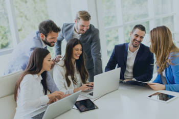 Six people sitting and standing around a white table in a bright office, working on laptops and tablets during a collaborative meeting.